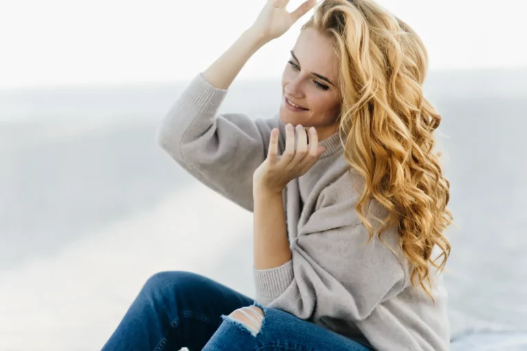 Jedna z uczennic szkoły fryzjerskiej Warszawa z długimi blond falowanymi włosami siedząca na zewnątrz.