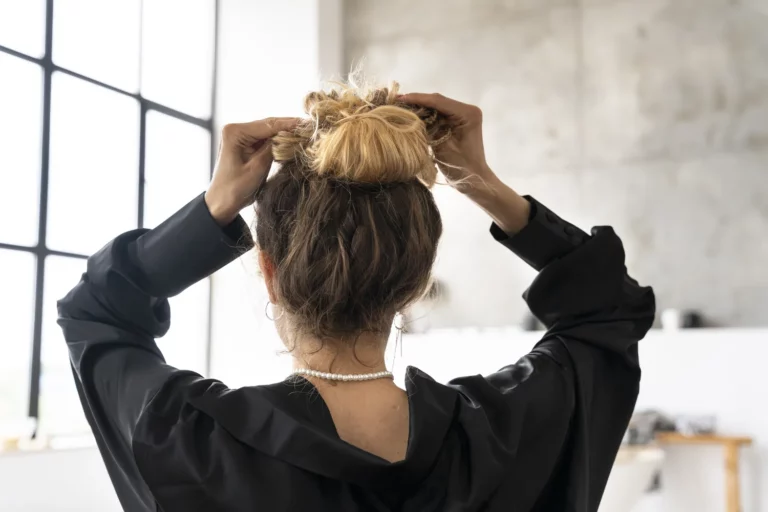 Widok od tyłu na kobietę we wnętrzu, poprawiającą luźny kok obiema rękami, ubraną w czarną bluzkę i naszyjnik z pereł, stojącą przy dużym oknie.