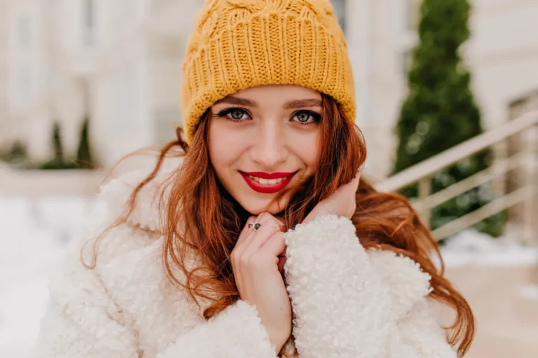 Uśmiechnięta kobieta w żółtej czapce zimowej z wyrazistą czerwoną szminką, efekt pracy rozwijanej przez szkoła wizażu Warszawa oraz kurs makijażu Warszawa