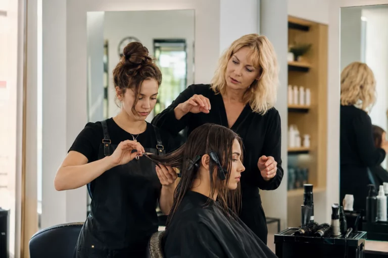 Kursantka ćwicząca technikę strzyżenia na modelce pod okiem instruktorki w nowoczesnym salonie fryzjerskim.