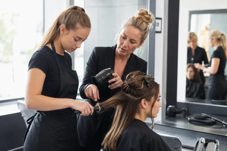 Instruktorka pokazuje kursantce technikę modelowania włosów na modelce w profesjonalnej sali szkoleniowej.